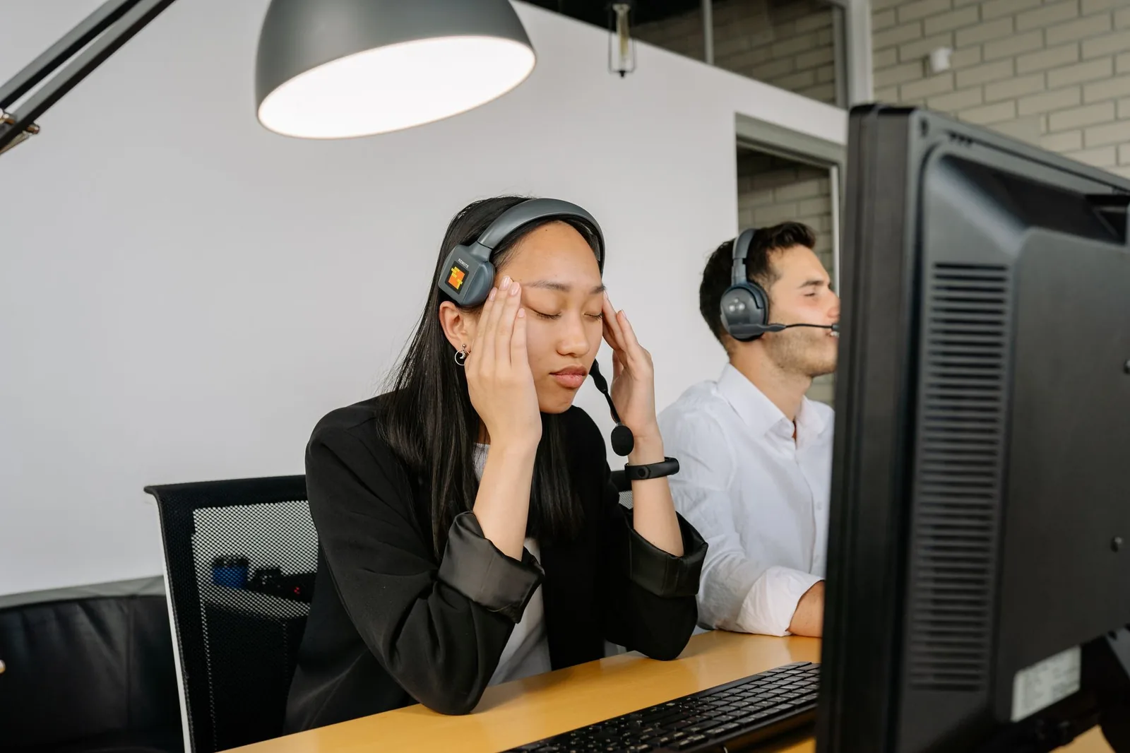 Asian woman at call center desk looking stressed, eyes closed, in office setting.