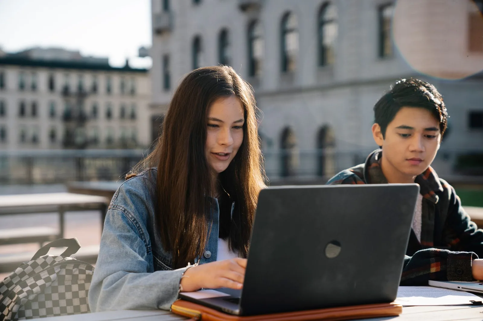 Two students collaborating on a laptop outdoors with a historic building in the background.