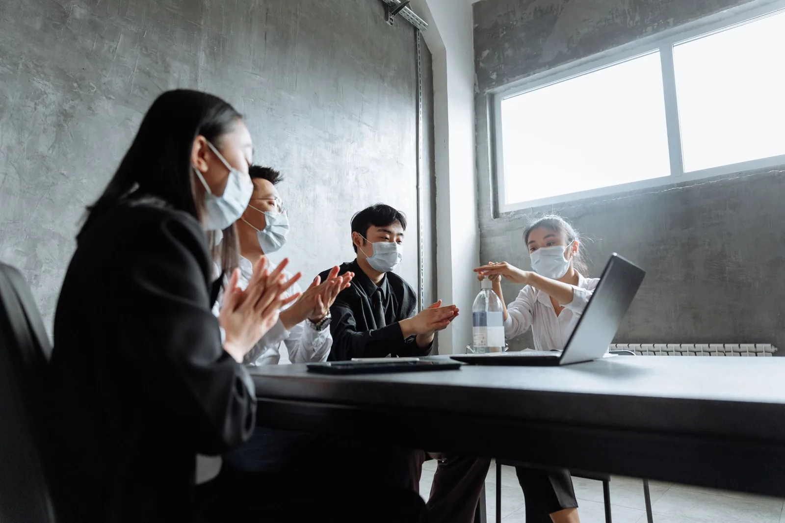Colleagues in an office setting use hand sanitizer, wearing masks during a meeting.