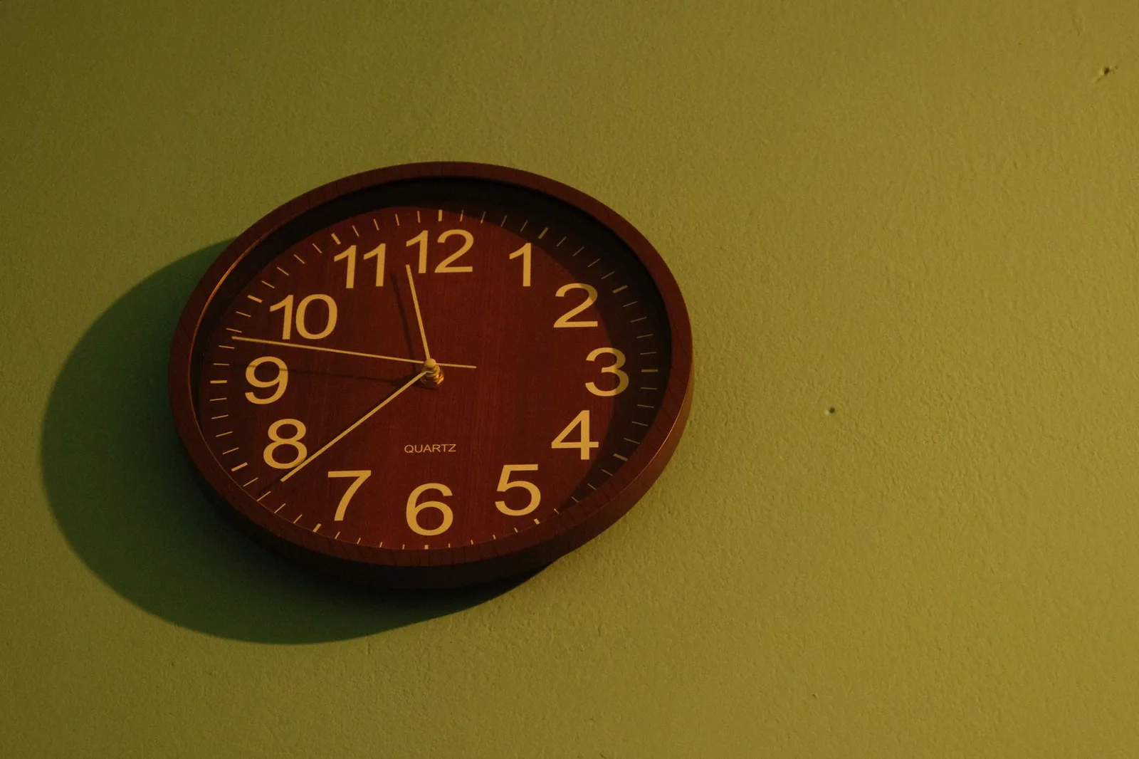 Brown wall clock with white numbers against a green wall, captured indoors.