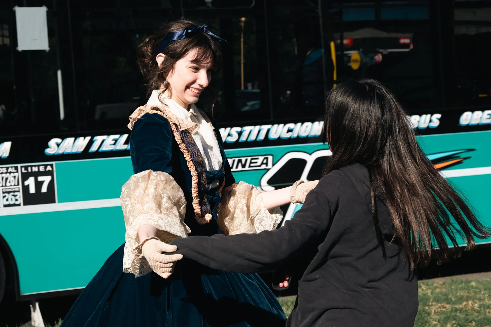 Two young women joyfully embrace in a street scene, one in vintage attire and a city bus in the background.